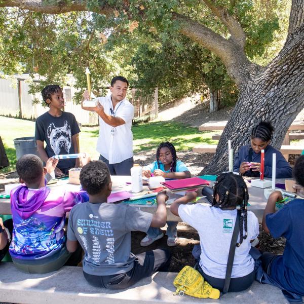 Students sitting outside while an instructor demonstrates a paper rocket.