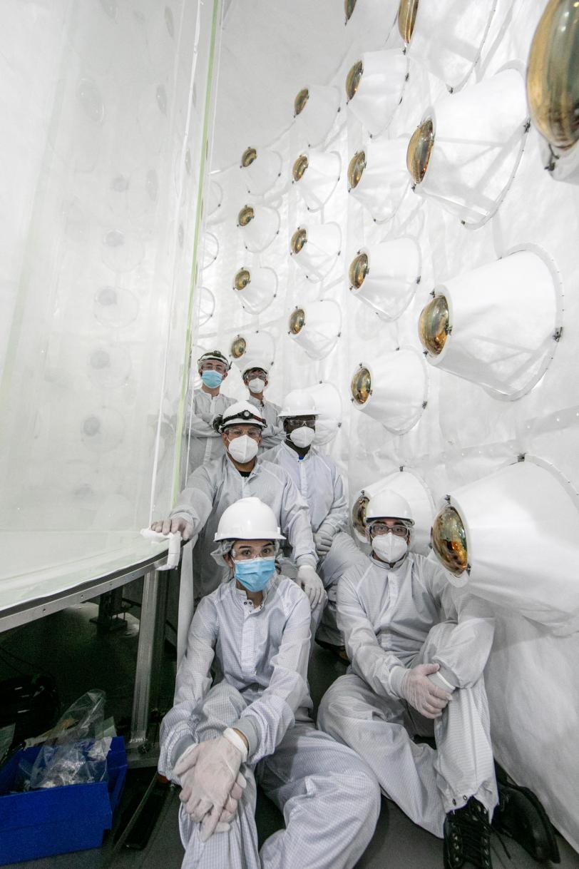 People in white jumpsuits sit between a white cylinder and a wall with mounted lenses.